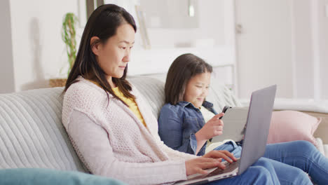 Video-of-happy-biracial-mother-and-daughter-sitting-on-sofa-with-laptop-and-tablet