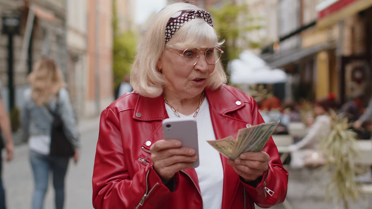 Happy senior woman counting money dollar cash use smartphone calculator app in urban city street