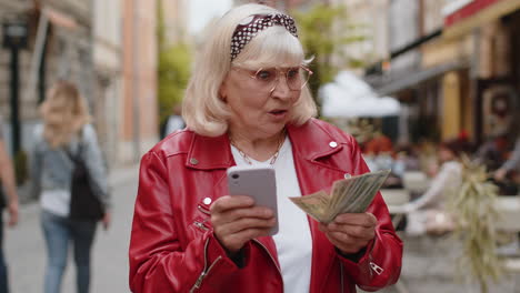 happy senior woman counting money dollar cash use smartphone calculator app in urban city street