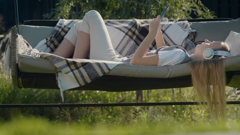 A-teen-girl-with-long-hair-listens-to-music-on-headphones,-lies-on-a-garden-swing-in-the-backyard-of-a-house.-4k-video