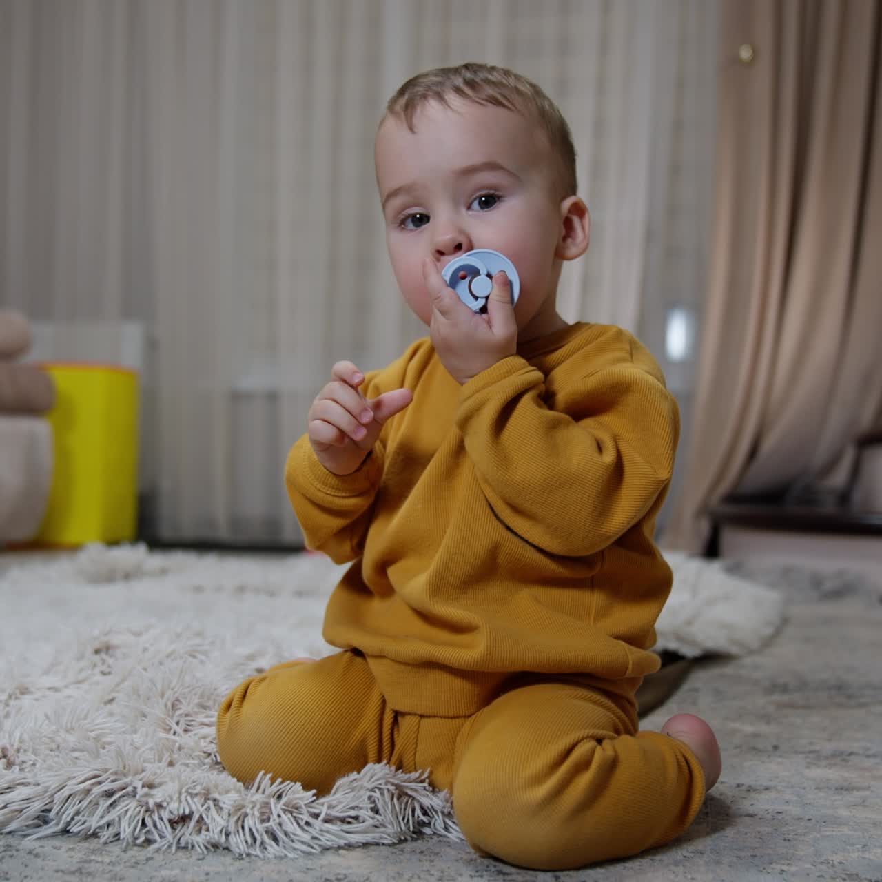 Pretty little baby sitting on the floor with pacifier in mouth. Kid looses the soother and picks it up and pulls to mouth