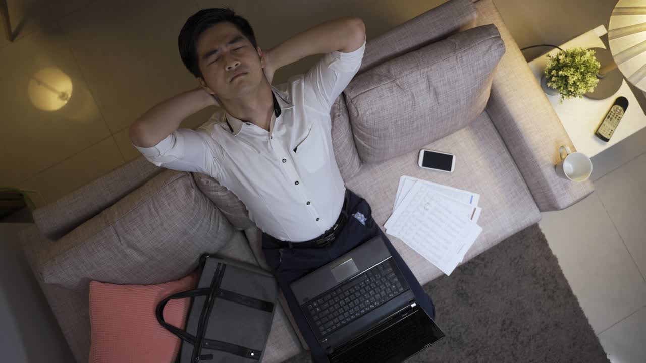 위에서 볼 때, 스트레스를 받는 아시아 남성 직원은 집에서 거실에서 노트북에서 늦게 일하면서 머리를 뒤로 던지고 위로 쳐다보고 있습니다.