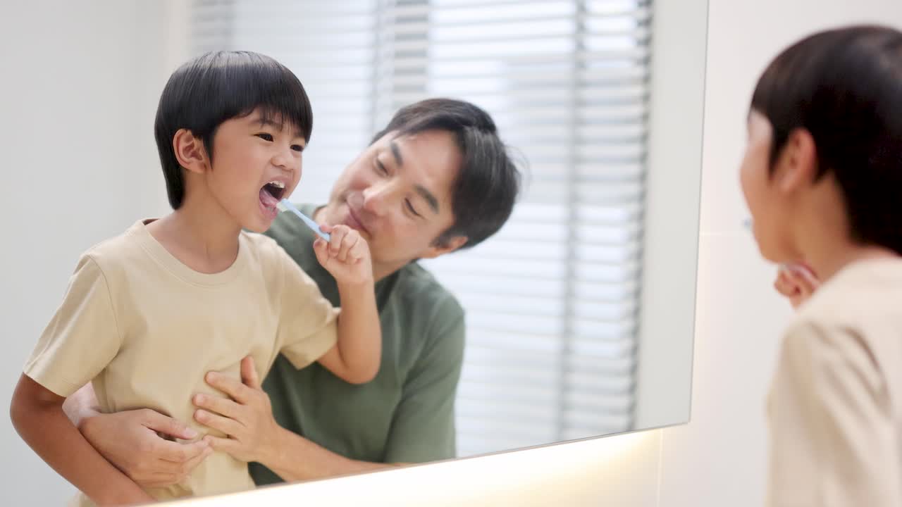 Father and son joyfully brush teeth together in a bright bathroom, smiling in the mirror