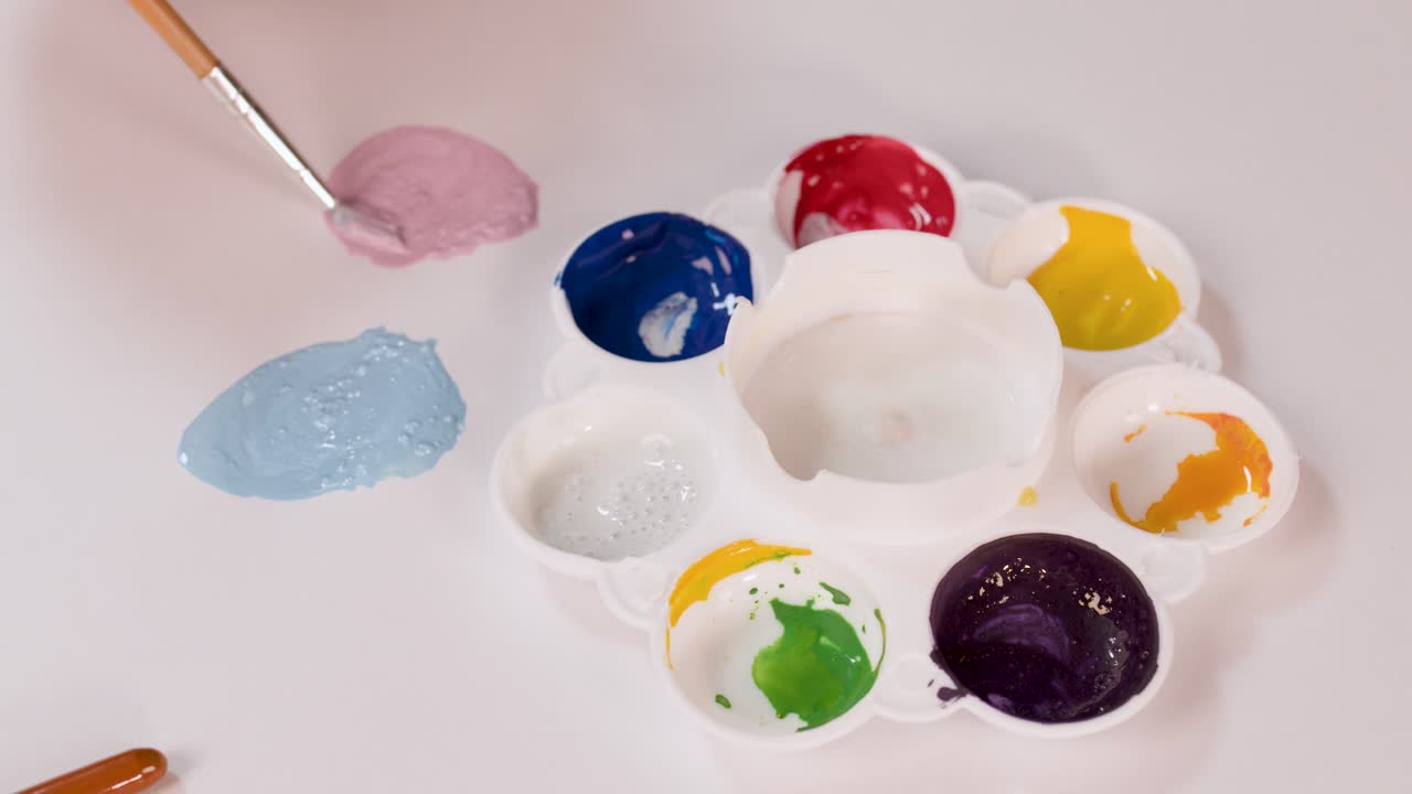 Artist hand mixes watercolor paints on palette, creating pastel shades under bright, even lighting