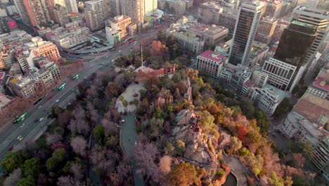 aerial orbit of santa lucia hill with autumnal trees, traffic in alameda avenue, santiago city buildings in back at sunset, chile