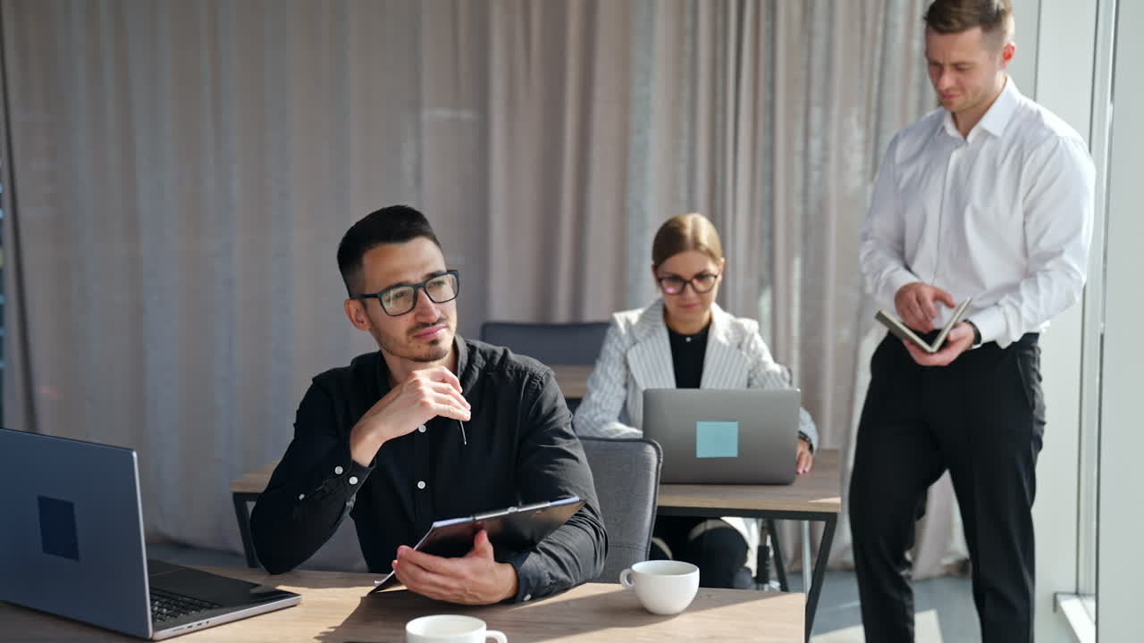 Pensive man in black shirt and glasses thinking over some work issues, ways of solving problem. Male manager talking to employees, checking data, controlling work process.
