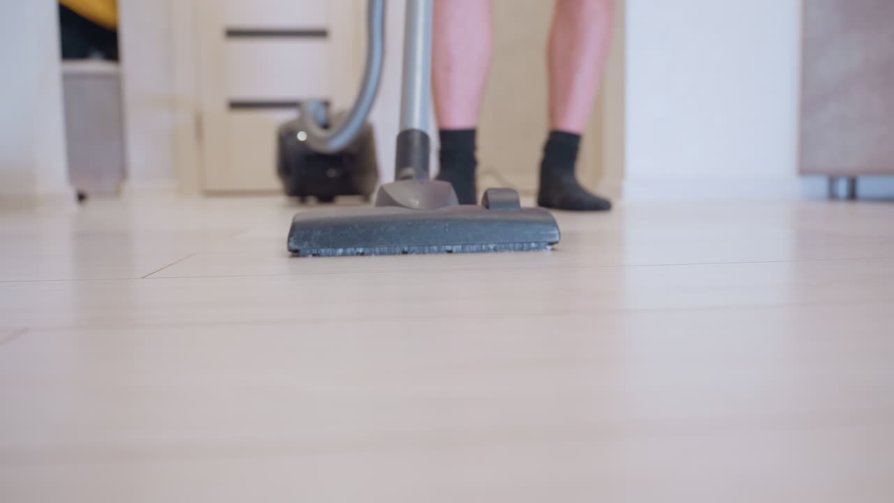 Low angle close up of vacuum cleaner head moving across light wooden floor with person in black socks visible in background, showing daily household cleaning activity and home hygiene