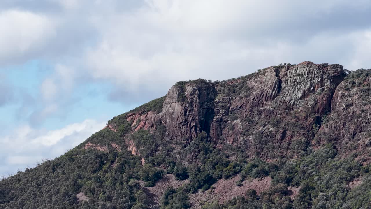 A slow horizontal pan reveals a rugged, tree-lined mountain under partly cloudy skies in Coonabarabran, New South Wales, with natural daylight and steady camera movement