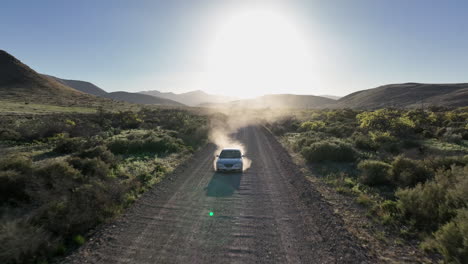 Aerial-shot-of-car-driving-on-dirt-road-in-Willcox,-Arizona,-drone-shot-with-mountains-in-the-background-and-dust-behind-the-car-into-the-sun