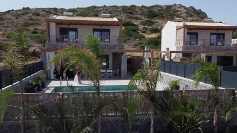 dark haired slim woman walks to a private pool and takes off the bathrobe and sits down in a swimming costume to the pool - villa in greece crete and palm trees in the foreground can be seen