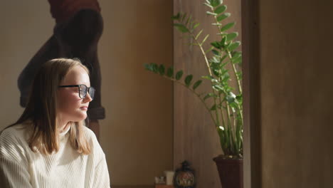 close up of decorative rattan wall light beside mural as pensive woman sits in cozy corner sipping tea, with spoon on table and potted flower nearby, soft ambient lighting completes calm scene