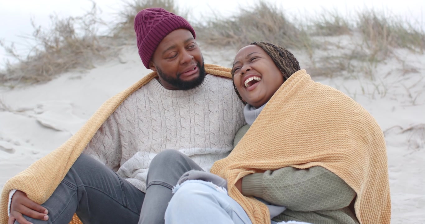 Coppia afroamericana seduta su una duna, l'uomo che avvolge una coperta color senape, in cerca di calore e vicinanza.