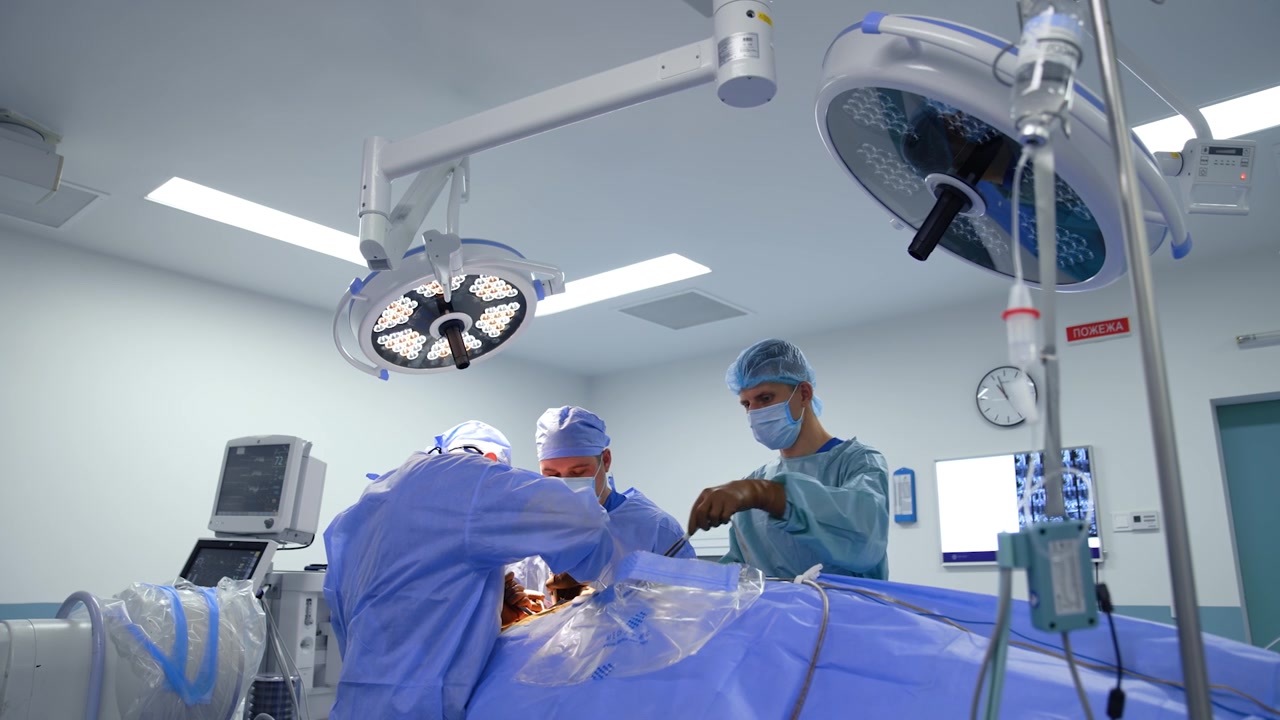 Team of three medics work at operating table. Male nurse takes a tool device and cleans it with sponge. Low angle view.