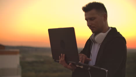 de pie al atardecer en el techo con una computadora portátil y una cerveza. un hombre con capucha trabaja divirtiéndose y contemplando la belleza de la vista de la ciudad desde una altura.