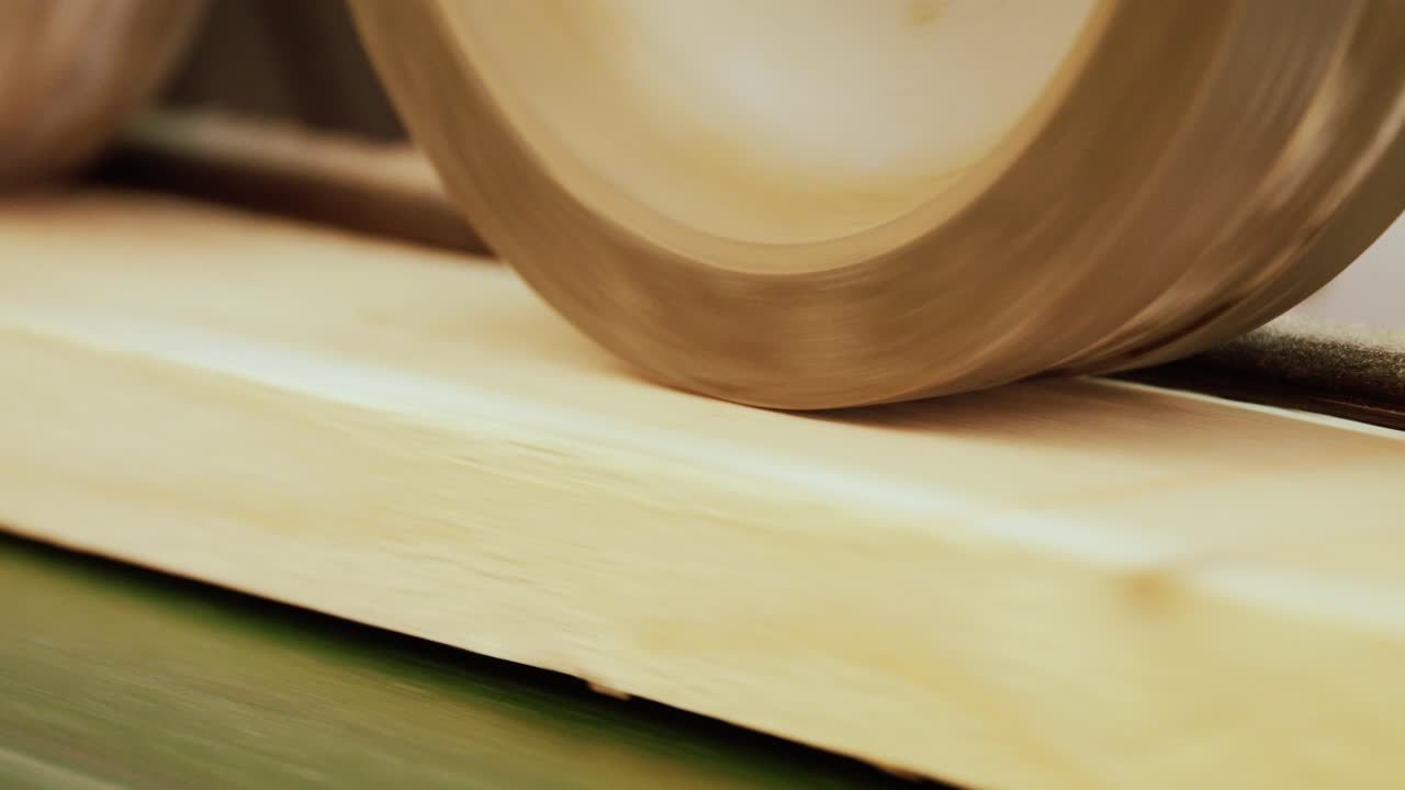 Close-up of an industrial woodworking machine. A large, spinning roller wheel presses down and feeds a wood plank forward, showcasing the power of the manufacturing process