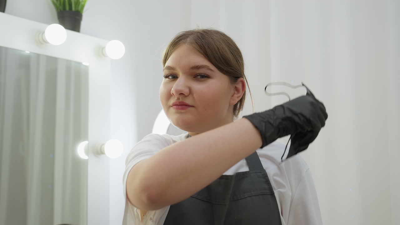 Beautician in black apron and gloves looks at camera while removing face mask and holding three cosmetic brushes. Bright salon interior with mirror and makeup tools visible in clean setup
