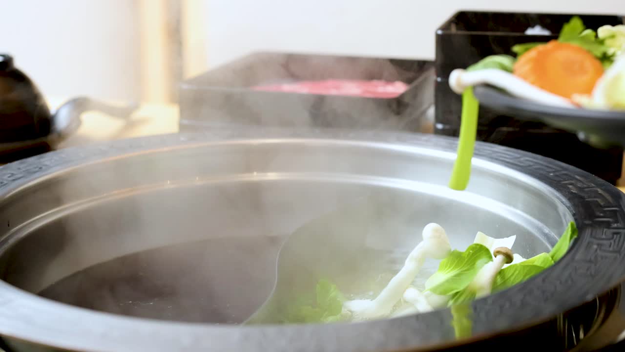 Hand places assorted fresh vegetables into steaming dual broth hotpot, bright lighting, close-up view