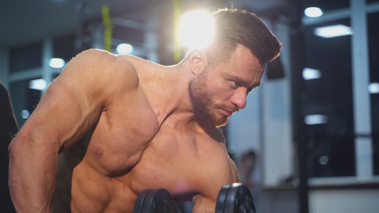 Fitness guy training indoors. Shirtless bodybuilder lifting heavy dumbbell. Muscular athlete exercising with a dumbbell in gym. Close-up.