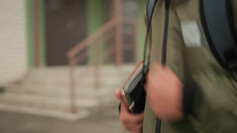 학생이 팔에 노트패드를 끼고 어깨에 백팩을 멘 채 달리는 모습 클로즈업, 대학 건물 철제 프레임과 녹지가 배경에 흐릿하게 보여 활기찬 학업 라이프스타일을 보여줍니다.