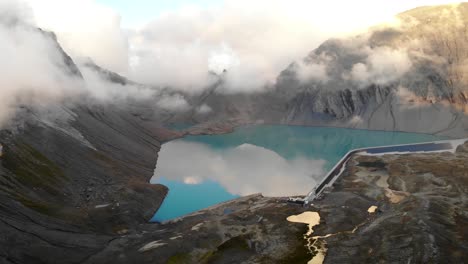 Aerial-view-of-lake-Muttsee-in-Glarus,-Switzerland-during-a-cloudy-sunset,-with-the-hydroelectric-dam-reservoir-full-of-water-and-solar-panels