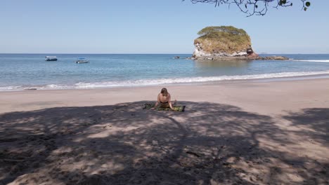 mujer joven en bikini sentada debajo de un árbol en una playa vacía en costa rica