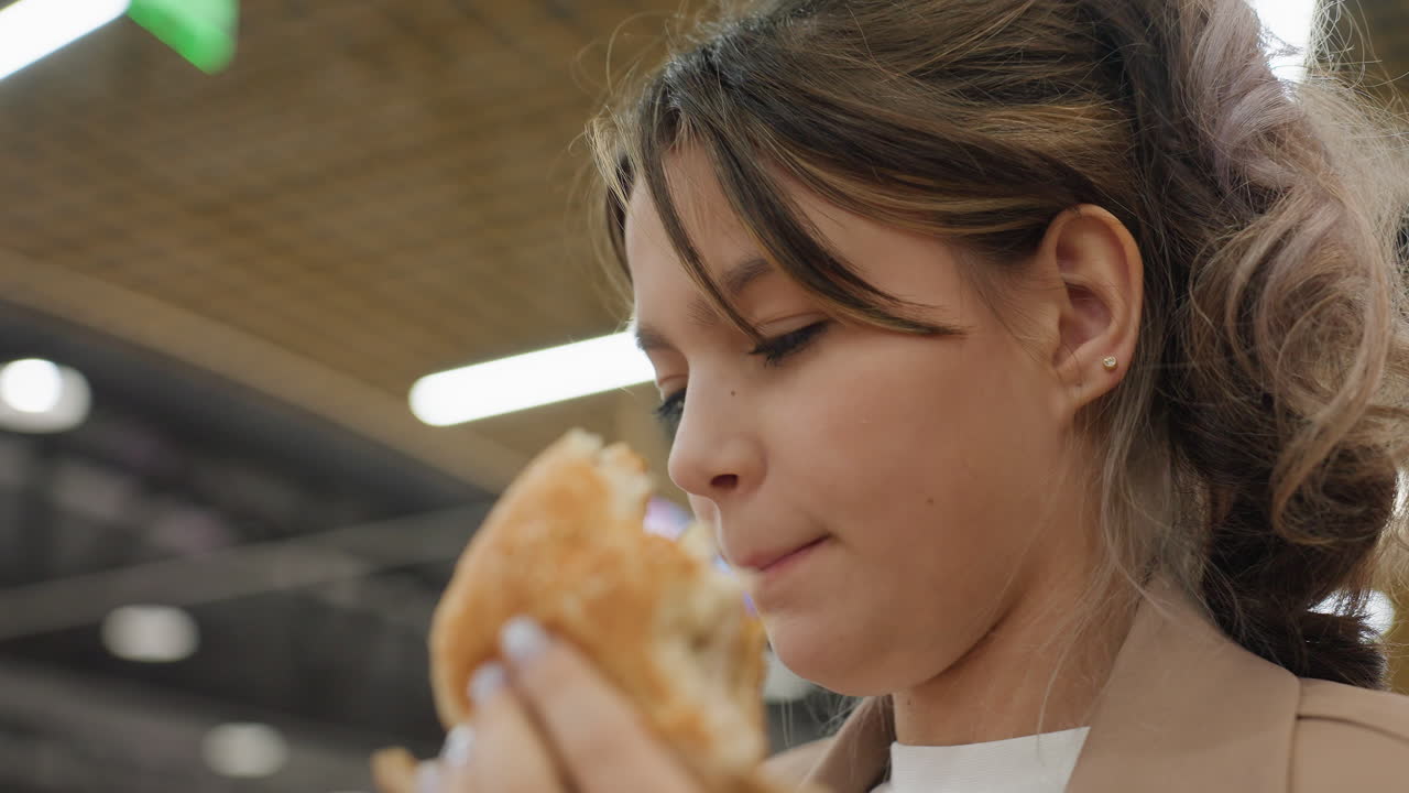 Mujer pensativa revisando su hamburguesa cuidadosamente en la animada zona de comedor del centro comercial; mujer pensativa que se detiene e inspecciona cuidadosamente su hamburguesa antes de darle un bocado en la concurrida zona de restauración del centro comercial.