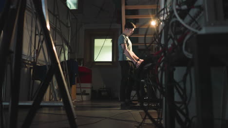 man in green t-shirt walks through dimly lit backstage technical room surrounded by equipment, metal ladders, tangled cables, and staircase while warm light illuminates notes on wall-mounted board nearby