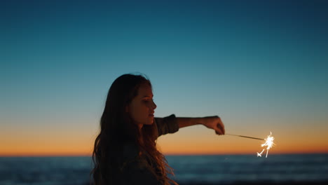 Frau-Spielt-Mit-Wunderkerzen-Am-Strand-Bei-Sonnenuntergang-Und-Genießt-Die-Silvesterfeier.-Mädchen-Feiert-Den-Unabhängigkeitstag-Und-Schwenkt-Wunderkerzen-Feuerwerk-Am-Meer