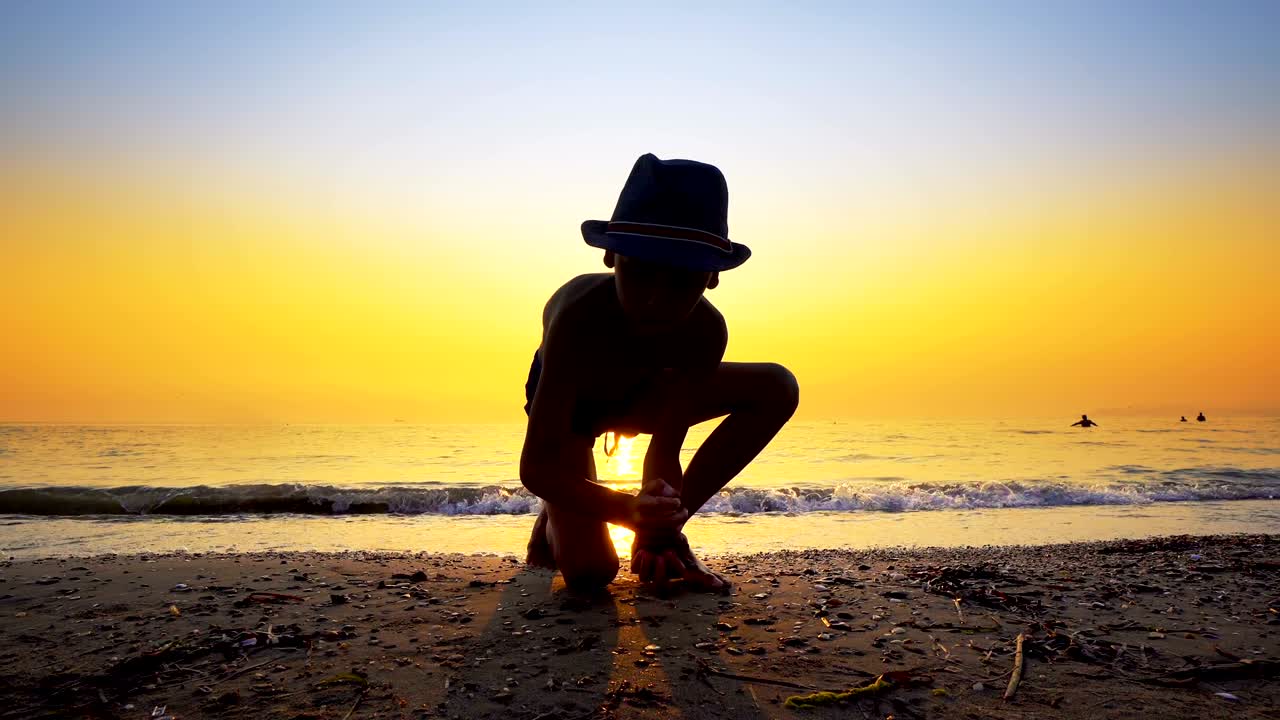 모자를 입은 소년의 실루은 바물 표면에서 돌을 던지고 점프합니다. 활기찬 오렌지색 하늘과 함께 여름 휴가 개념