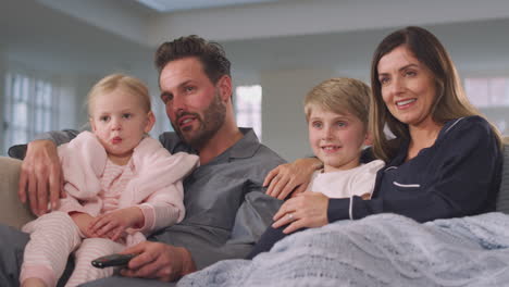family in pyjamas sitting on sofa watching tv together