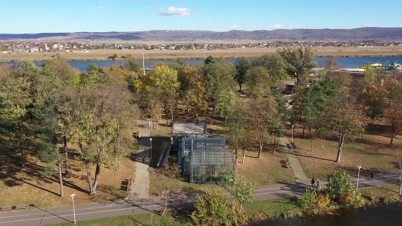 An aerial drone orbits a modern glass building, likely a greenhouse or pavilion, in a scenic riverside park. Bacau. People stroll along the paths among the colorful autumn trees