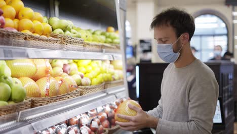 man in mask takes a fresh citrus from the food shelf. shopper choosing pummelo at grocery store