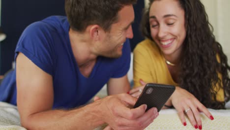 una feliz pareja caucásica acostada en la cama y usando un teléfono inteligente