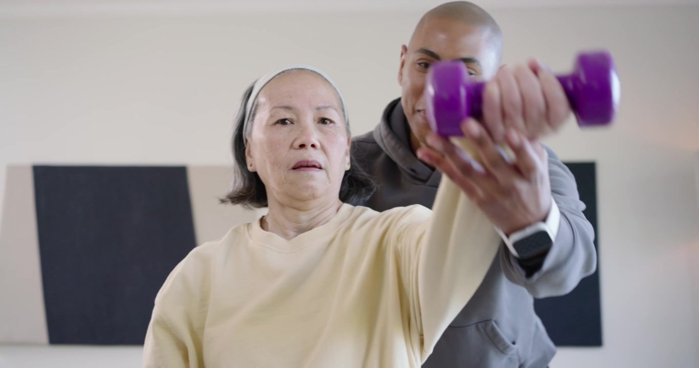 Allenatore che si china, guida un'anziana donna cinese con un peso viola, mentre solleva il braccio per la riabilitazione in clinica