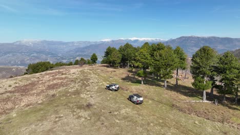 Panoramaaufnahme-Von-Zwei-Geländefahrzeugen,-Die-Auf-Einem-Hügel-Fahren,-Mit-Wald-Und-Schneebedeckten-Bergen-Im-Hintergrund