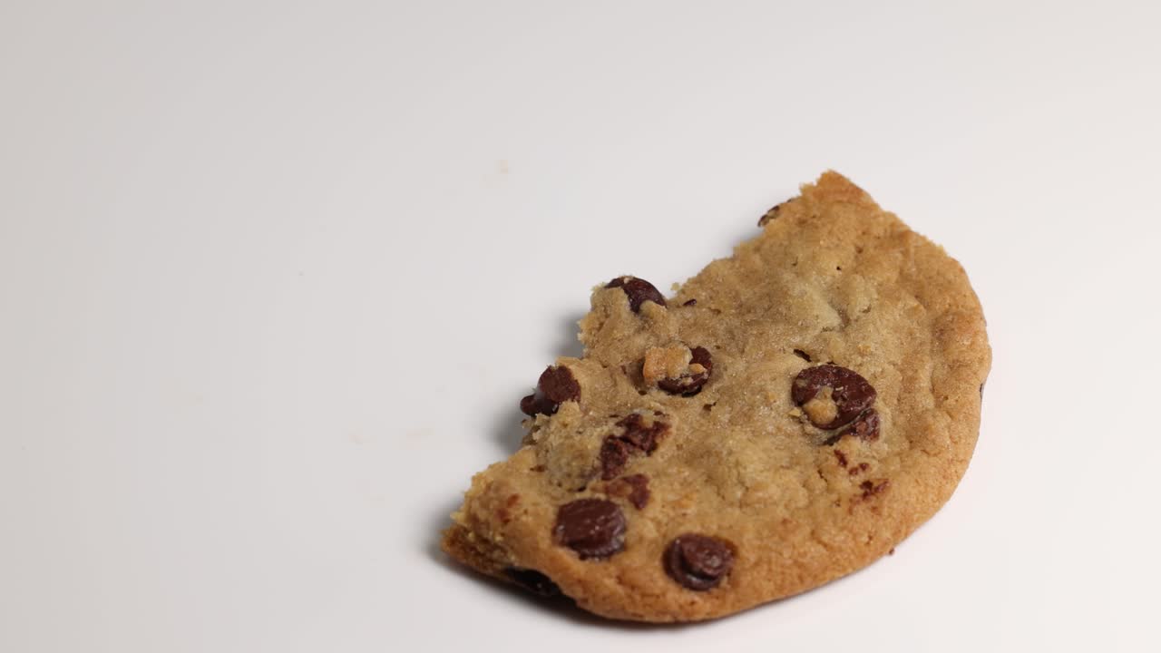 A hand breaks and picks up a chocolate chip cookie on a white surface under soft lighting