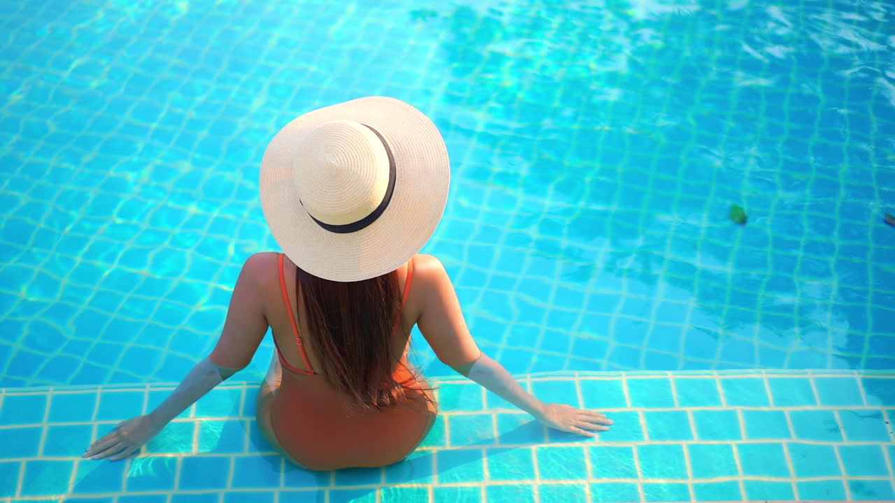With her back to the camera, a young fit woman sits o the shallow step of a swimming pool