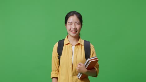 asian woman student with a backpack and some books standing and smiling to camera in the green screen background studio