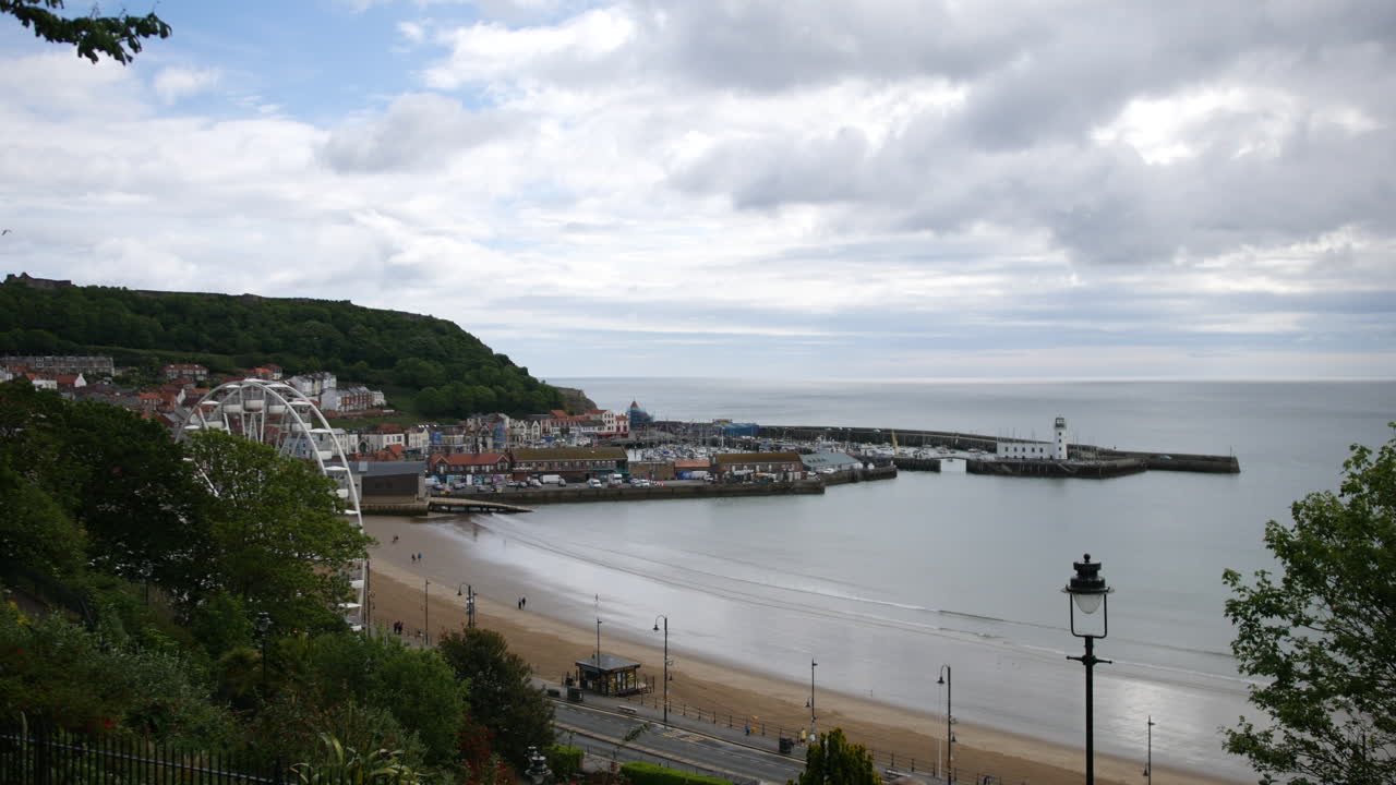 A coastal view of Scarborough, North Yorkshire in England showing a sandy beach, ferris wheel, lighthouse, and marina curving around the calm bay