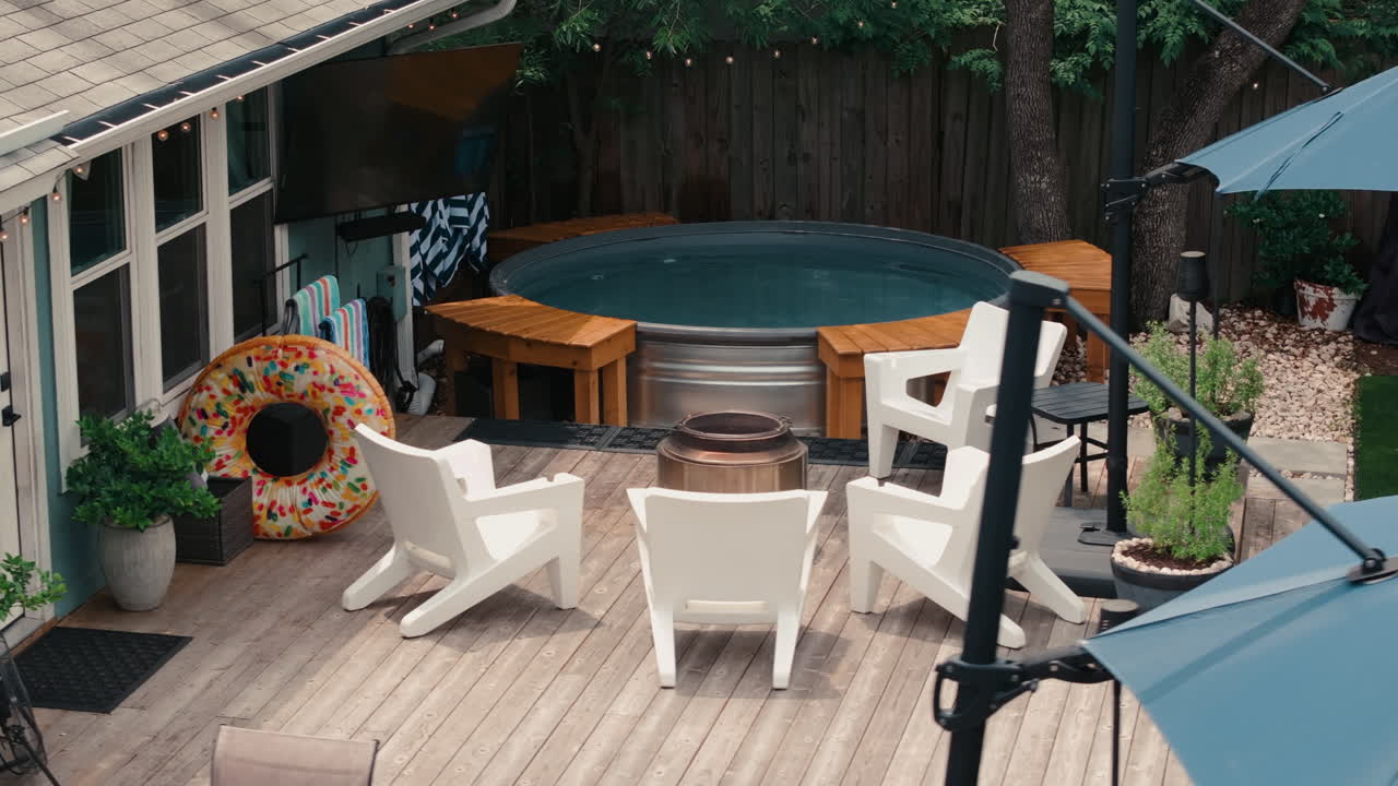 Elevated drone shot of a backyard deck with a hot tub, white Adirondack chairs, donut float, and outdoor TV under string lights. Perfect for outdoor living ads, lifestyle, and home content