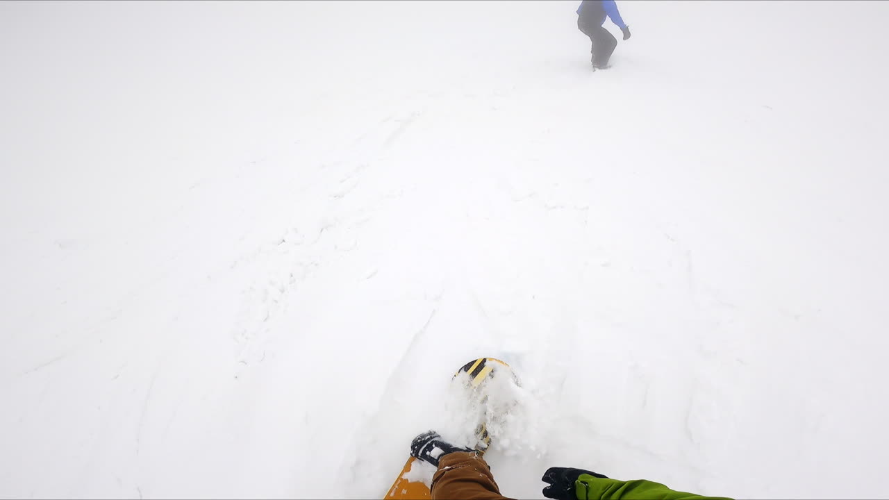 First person's footage of own feet and snowboard during the descend. Fellow snowboarder rides ahead.