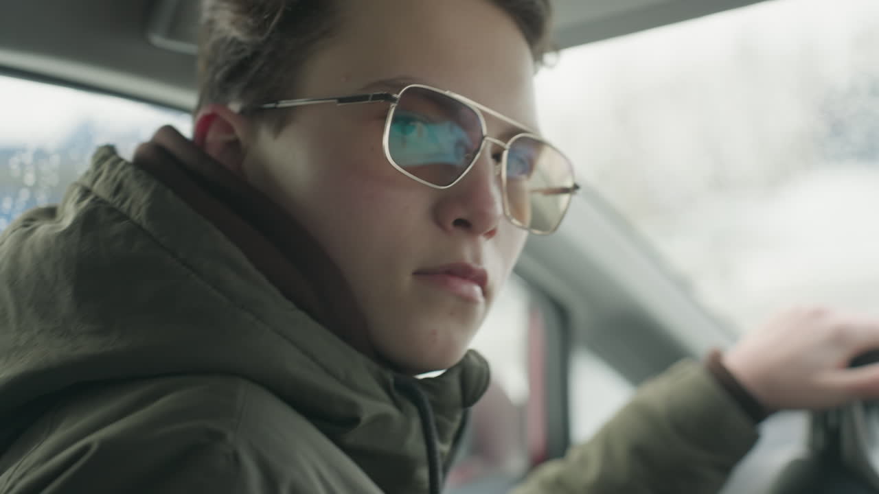 close up of man wearing glasses driving vehicle as he turns to look at camera with soft focus snowy urban background visible through window and windshield on cold winter day
