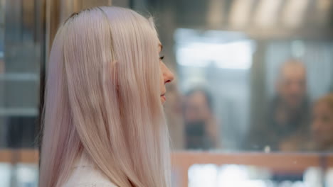 thoughtful pedestrian looking through transparent elevator glass at reflected mall scene, profile with pastel hair and sweater, blurred figures in mirrored cabin, ambient light bokeh