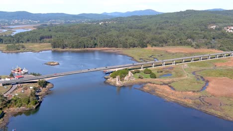 Bridge-with-cars-circulating-over-the-Ulla-River-and-the-ruins-of-the-towers-of-the-West-Viking-landing-zone,-sunny-afternoon,-drone-shooting-traveling-forward-zenithal-1