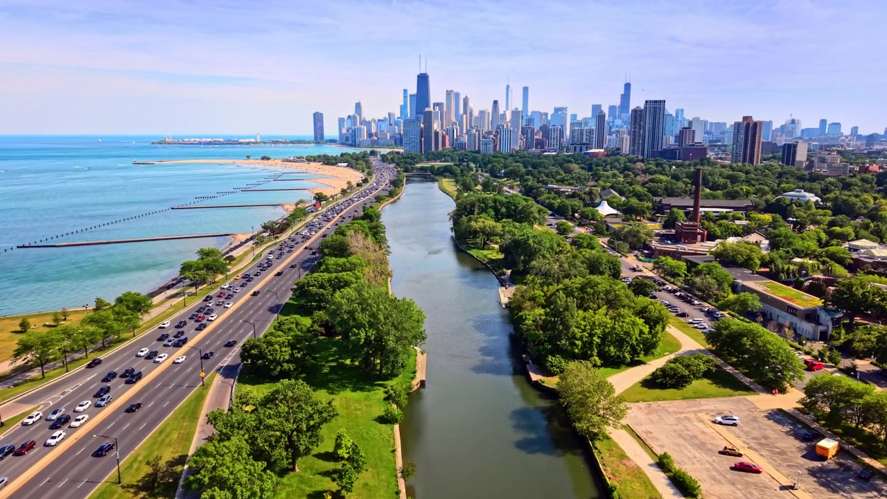 Coastal highway alongside park and river, Chicago. Drone view of busy traffic, green trees, water canal and distant skyline.