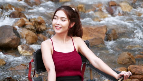 mujer asiática de rojo sentada en una silla con grandes rocas y agua salpicando lentamente para relajarse y ser feliz en las vacaciones de verano