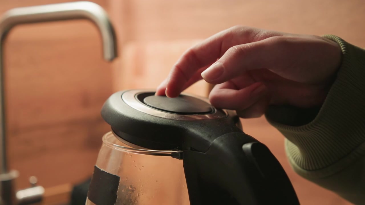 Right hand steadies black kettle lid while clear glass carafe fills with hot water on wooden kitchen counter, with steam, droplets, warm light, and domestic routine in view, with kettle, lid, hand