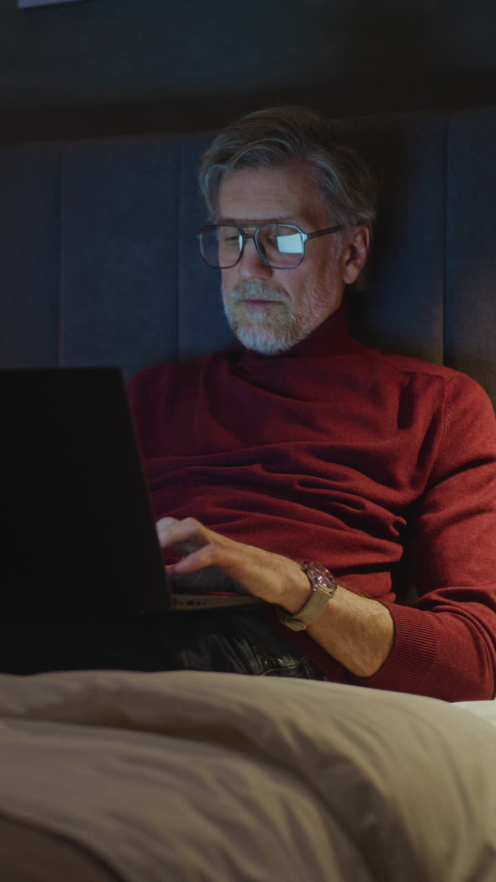 Elegant Mature Man Wearing Glasses Uses Laptop While Sitting On Bed (Stock Footage)