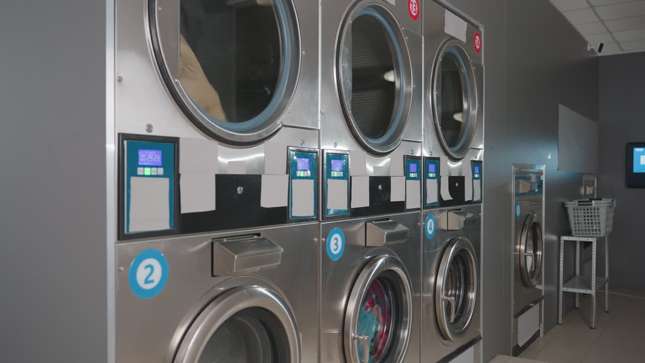 Row of steel washing machines spinning with clothes inside, digital timers glowing, clean modern laundry facility, baskets on stand, active wash cycle visible through transparent glass doors