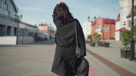 back view of a lady with brown hair in a black coat adjusting her head, showcasing three rings on her left hand on a sunny urban street with residential building with background